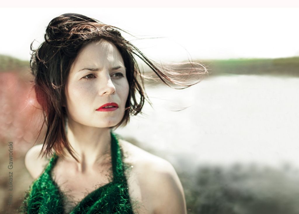 na pierwszym planie Magdalena Kumorek.  Wpatrzona w dal. Tle jezioro. Rozwiane rude włosy. Zielone sukienka odsłaniająca ramiona. porcelanowa, jasna skora.